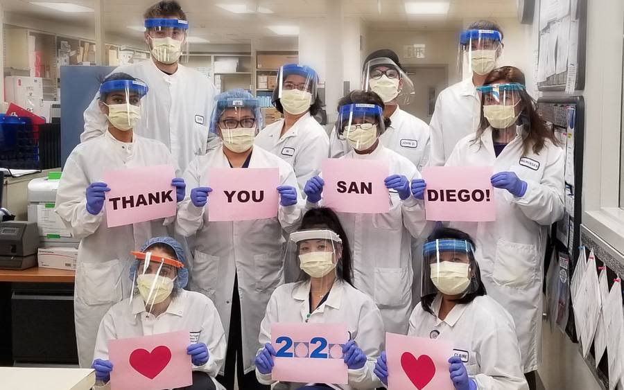 During the COVID pandemic, Scripps laboratory staff wear full PPE and small signs that together say 
